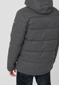 Giacca imbottita grigia con cappuccio, caratterizzata da una trapuntatura orizzontale e da un logo applicato sul braccio sinistro. Tessuto testurizzato e colore uniforme.