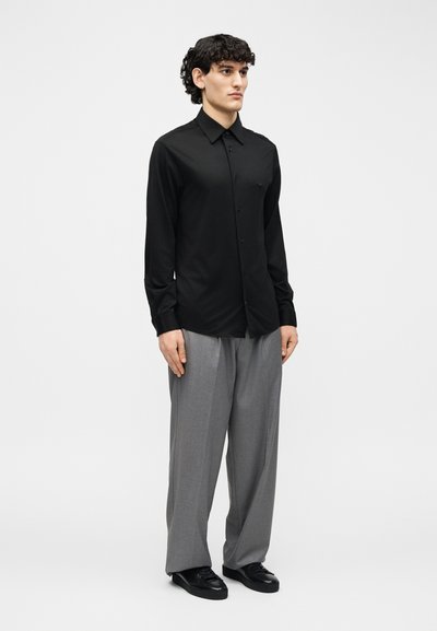 Άνδρας με μαύρο κουμπωτό πουκάμισο, άνετο γκρι παντελόνι και μαύρα παπούτσια, στέκεται με τα χέρια χαλαρά στα πλευρά, σε λευκό φόντο.