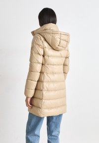 Kvinna med mörkt hår iklädd en beige quiltad knä-lång jacka med huva och ljusblå jeans, stående mot en enkel bakgrund.