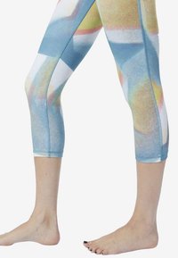 Nogi ubrane w 3/4 legginsy z abstrakcyjnymi pastelowymi wzorami w odcieniach niebieskiego, żółtego, różowego i białego, stojące boso na białym tle.