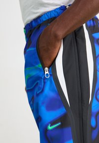 Nærbillede af en hånd i den lynlåste lomme på blå, sorte og hvide Nike sportsbukser med et neon-grønt logo.