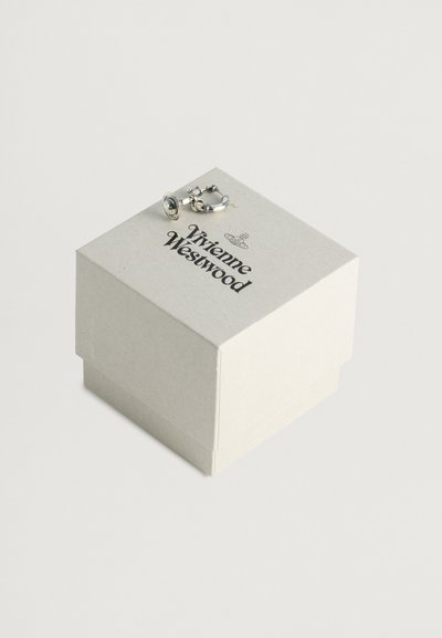 Kvadrāta formas, gaiši brūna kartona kaste ar melnu tekstu "Vivienne Westwood". Uz augšu novietota sudraba auskaru, kas parāda gredzena dizainu.