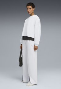 Felpa bianca cropped con vestibilità ampia, abbinata a pantaloni bianchi a gamba larga. Presenta un logo sottile e dettagli testurizzati. Tiene una borsa nera.