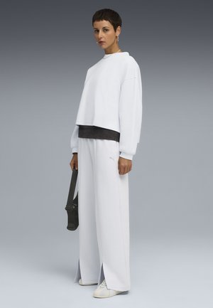Hvid cropped sweatshirt med løs pasform, parret med brede hvide bukser. Har et diskret logo og teksturerede detaljer. Holder en sort taske.