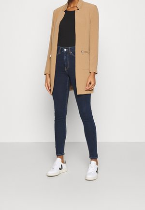 Blazer lungo beige con design sartoriale, due tasche frontali e cerniere argentate; abbinato a jeans skinny blu scuro e sneakers bianche.