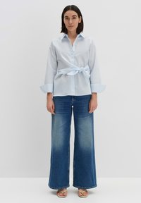 Camicetta azzurro chiaro a righe con una cintura in vita; abbinata a jeans a gamba larga di un blu sbiadito. I sandali completano l'outfit.