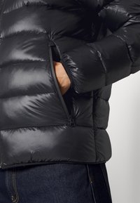 Chaqueta acolchada negra con un acabado brillante, que presenta un bolsillo lateral con cremallera. El material es suave y aislante, con un diseño ajustado.