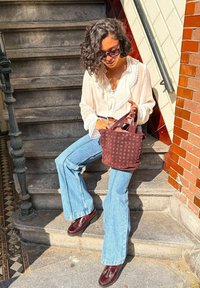 Bruine geperforeerde handtas met korte handvatten, witte blouse met kantafwerking, lichtblauwe flared jeans en bordeauxrode schoenen op stenen trappen.