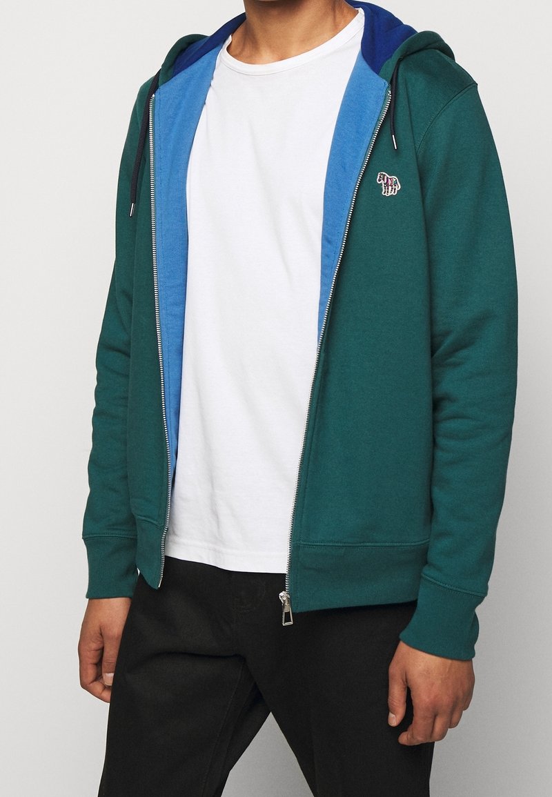 Grüne Zip-Hoodie mit einer blauen Innenschicht, ausgestattet mit einem kleinen gestickten Logo auf der Brust sowie gerippte Bündchen und Saum.