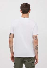 Biała bawełniana koszulka z okrągłym dekoltem i krótkimi rękawami, z widocznym jednolitym wzorem na plecach. Uzupełniona widocznymi tatuażami na ramionach.