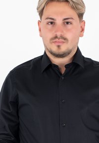 Camicia nera con bottoni, caratterizzata da una texture liscia, con colletto a punta e maniche lunghe, esposta su uno sfondo neutro.