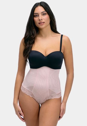 Femme aux longs cheveux foncés portant un soutien-gorge noir sans bretelles et une culotte gainante taille haute rose clair avec bordure en dentelle, debout devant un fond gris.