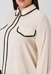 Frau in einer cremefarbenen Bluse mit schwarzem Besatz am Kragen, der Knopfleiste und zwei Brusttaschen, gezeigt von Hals bis zur Mitte des Oberkörpers.