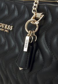 Borsa a tracolla nera trapuntata con una texture lucida, hardware dorato e un accento di portachiavi con un'etichetta. Logo visibile sul davanti.