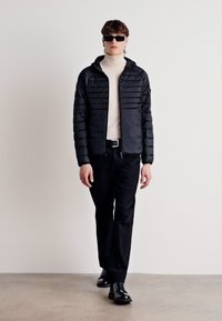 Svart puffjacka med horisontell quiltad design, dragkedjestängning och huva. Burit över en ljus polotröja, matchad med svarta byxor och skor.