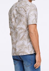 Chemise à manches courtes en tissu beige avec un motif de feuilles de palmier blanches. Dotée d'un col classique et d'un ourlet droit. Portée avec un jean bleu.