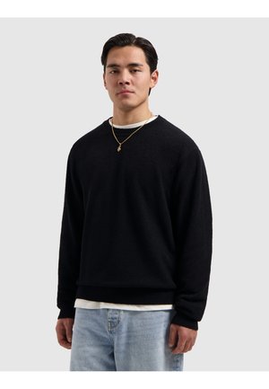 Junger Mann mit dunklen Haaren, der einen schwarzen Pullover, hellblaue Jeans, ein weißes Unterhemd und eine goldene Halskette trägt, steht vor einem schlichten Hintergrund.