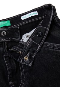 Vaqueros de mezclilla negros con cremallera parcialmente abierta, botón y cierres a presión, con etiquetas de la marca verdes y blancas en el interior de la pretina.