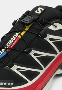 Schwarzer Trailrunning-Schuh aus Gore-Tex-Material mit gemustertem Obermaterial, elastischem Schnürsystem und roten, weißen sowie gelben Akzenten.