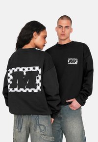 Schwarzer Sweatshirt mit großem schwarzen "M"-Patch und karierter Muster auf dem Rücken; die Vorderseite zeigt ein kleines Etikett mit dem Text "MCT PX".