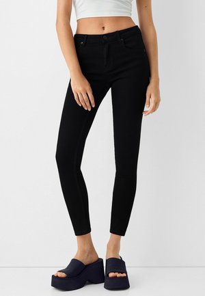 Žena v černých skinny džínech a černých platformových sandálech, s bílým krátkým topem, postávající na jednolitém pozadí.