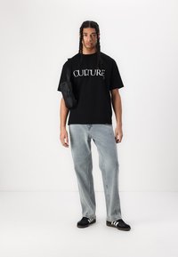 Zwarte T-shirt met korte mouwen met de tekst "CULTURE", gecombineerd met lichtblauwe spijkerbroek en zwarte sneakers met witte strepen. Kleine tas bevestigd.