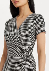 Czarna i biała sukienka w wzór houndstooth z dekoltem w kształcie litery V, krótkimi rękawami i fasonem kopertowym, podkreślona detalem w postaci węzła po boku. Miękka, teksturowana tkanina.