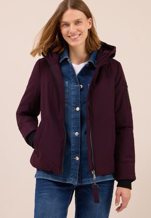 Veste en duvet bordeaux avec fermeture éclair à l'avant, capuche et poches latérales, superposée sur une chemise en denim. Présente une texture lisse et des poignets ajustables.