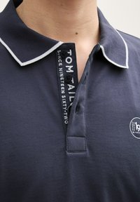 Tmavě modrá polo košile s bílým límečkem a knoflíkovými akcenty. Obsahuje značenou pásku na krku a logo na hrudi. Hladký textilní povrch.