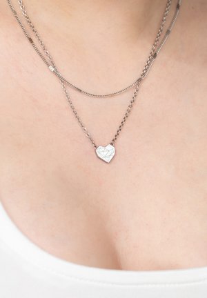 Silberne Doppelketten-Halskette mit einem geometrischen Herzanhänger, getragen auf nackter Haut über einem weißen Oberteil.