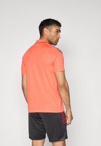 Polo homme corail avec col, manches courtes et trois rayures navy sur les épaules. Associé à un short gris foncé.
