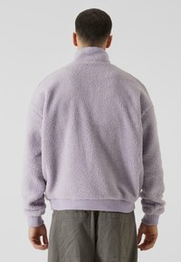 Giacca in pile viola con colletto alto, caratterizzata da una superficie texturizzata e soffice, polsini a costine e un taglio alla vita.