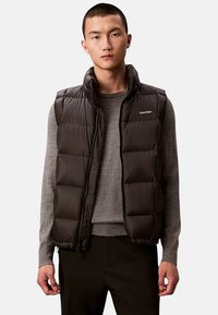 Czarna ocieplana kamizelka puchowa z wysokim kołnierzem, zamkiem błyskawicznym i logo na piersi. Posiada pikowane sekcje i gładką fakturę tkaniny.