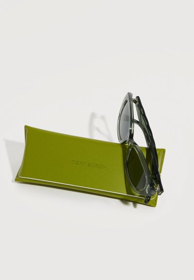 Γυαλιά ηλίου με διάφανη σκελετό που ακουμπούν σε μία θήκη από δέρμα σε φωτεινό πράσινο χρώμα της Tory Burch, σε λευκό φόντο.