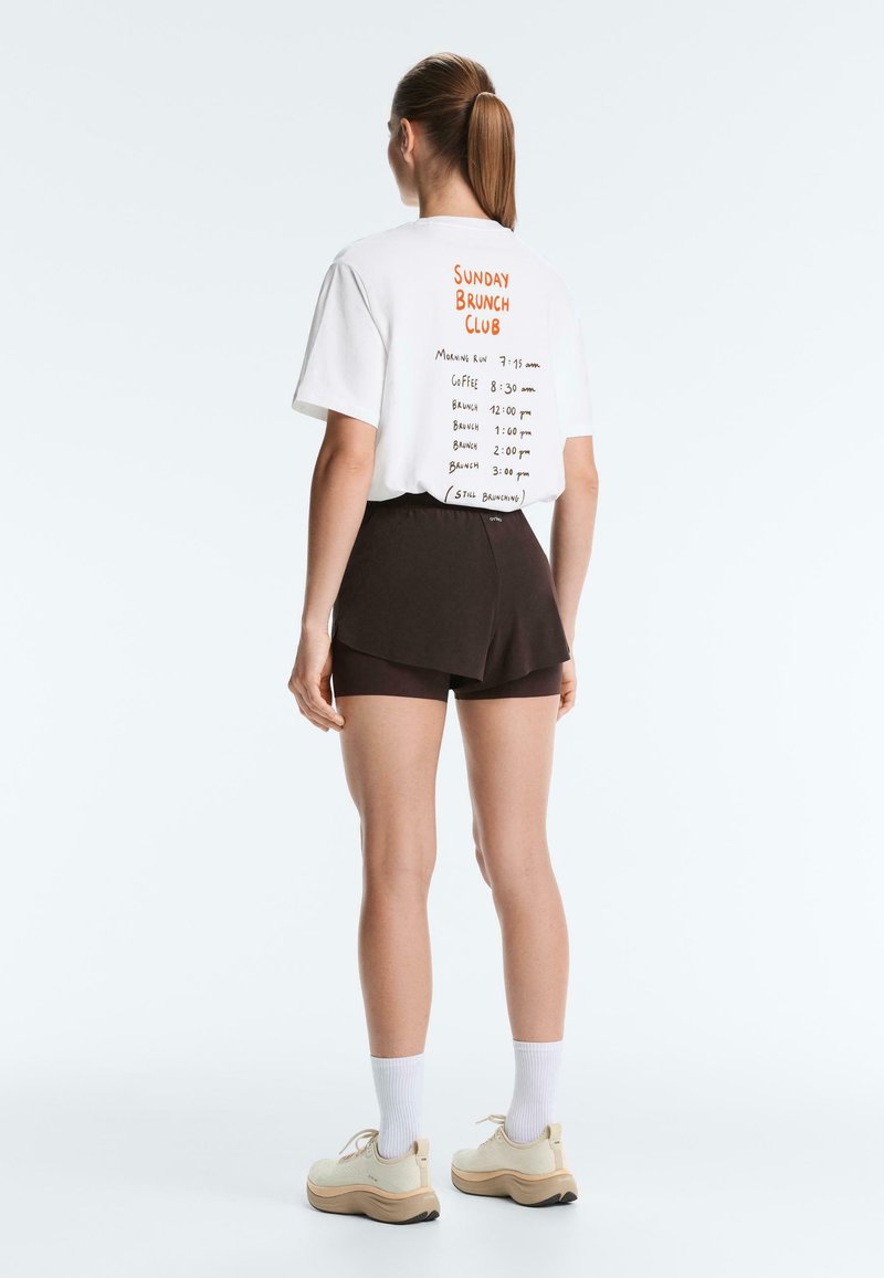 Vrouw met een paardenstaart, een witte "Sunday Brunch Club" t-shirt, zwarte korte broek, witte sokken en beige sneakers, staand met de rug naar een witte achtergrond.