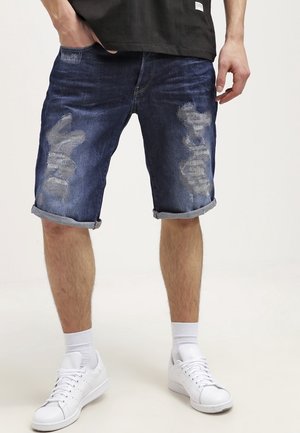 Szorty dżinsowe w ciemnoniebieskim kolorze z przetarciami, z podwiniętymi nogawkami i klasycznym pięciokomorowym fasonem. Połączone z białymi trampkami.