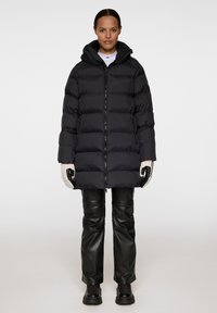 Svart pufferjacka med huva, med horisontella kanaler, framdragkedja och sidofickor, i kombination med svarta läderbyxor och handskar.