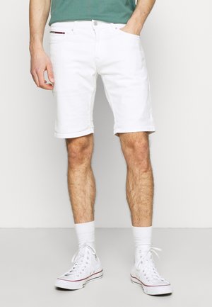 Homme portant un short blanc jusqu'aux genoux, des baskets blanches montantes, des chaussettes blanches et une chemise verte, les mains dans les poches.