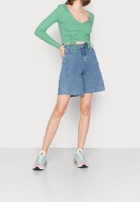 Grünes, geripptes Langarmoberteil mit Kordelzugdetails, kombiniert mit hochgeschnittenen blauen Denim-Shorts und mehrfarbigen Turnschuhen auf neutralem Hintergrund.