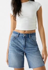 Donna con una maglietta bianca corta e pantaloni shorts di jeans blu a vita alta con orli sfilacciati, in piedi contro uno sfondo neutro.