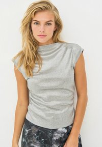 Plavuša s raspuštenom kosom nosi srebrni rebrasti top bez rukava i tamne hlače s cvjetnim uzorkom nasuprot jednostavne bijele pozadine.