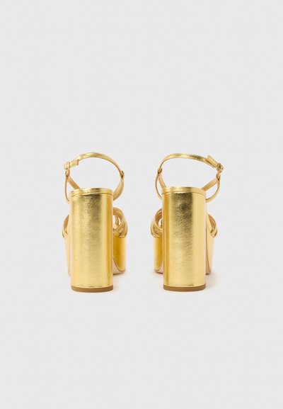 Ζευγάρι μεταλλικές χρυσές σανδάλες με ψηλά τακούνια, λουράκια στον αστράγαλο και χοντρά τακούνια, βλέποντας από πίσω σε απλό ανοιχτό φόντο.