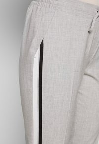 Pantalon gris clair avec taille élastique et poches latérales, présentant des rayures verticales noires et blanches le long de la couture extérieure.