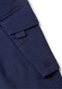 Námořnická modrá látka s hladkým povrchem, se šikmou kapsou a vyšitou logem v pravém dolním rohu.