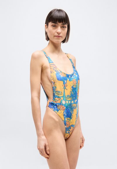 Γυναίκα με κοντά σκούρα μαλλιά, φοράει πολύχρωμο, ψηλόμεσο μαγιό με αφηρημένα πορτοκαλί και μπλε σχέδια, στέκεται μπροστά σε απλό γκρι φόντο.