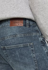 Vaqueros de mezclilla en azul oscuro con un acabado texturizado, con una etiqueta de cuero marrón que dice "REDEFINED REBEL" en la cinturilla.