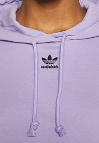 Helle lila Kapuzenpullover mit Kordelzug, gerippten Bündchen und einem schwarzen Adidas-Logo mit drei Streifen darüber. Aus weichem Baumwollmaterial.
