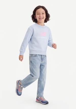 Niño sonriendo y caminando, vistiendo una sudadera azul claro de New Balance, jeans claros y zapatillas rosas y azules, con un fondo blanco.