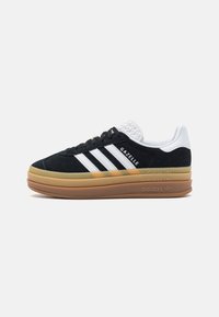 Tenisky Adidas Gazelle s černým semišovým svrškem, bílými koženými doplňky, třemi bílými pruhy a podrážkou z gumové pryže. Texturovaný detail na patě.
