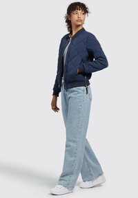Chaqueta bomber acolchada azul marino con cremallera al frente, puños y dobladillo acanalados; llevada sobre una blusa gris, combinada con vaqueros de pierna ancha color azul claro y zapatillas blancas.